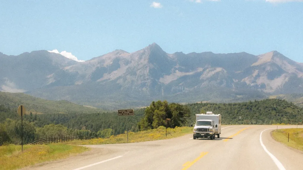 An RV drives down a road with large mountains in the background. 