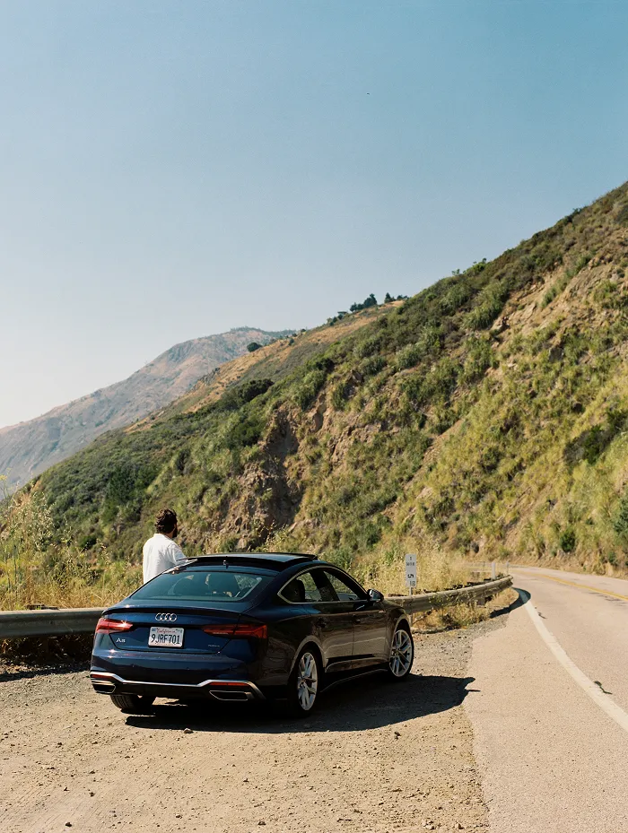 A man stands next to his car on the side of a mountain road.