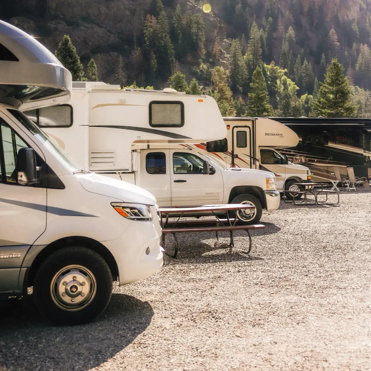 4 motorhomes parked in gravel sports with picnic tables in between and the forest in the background.