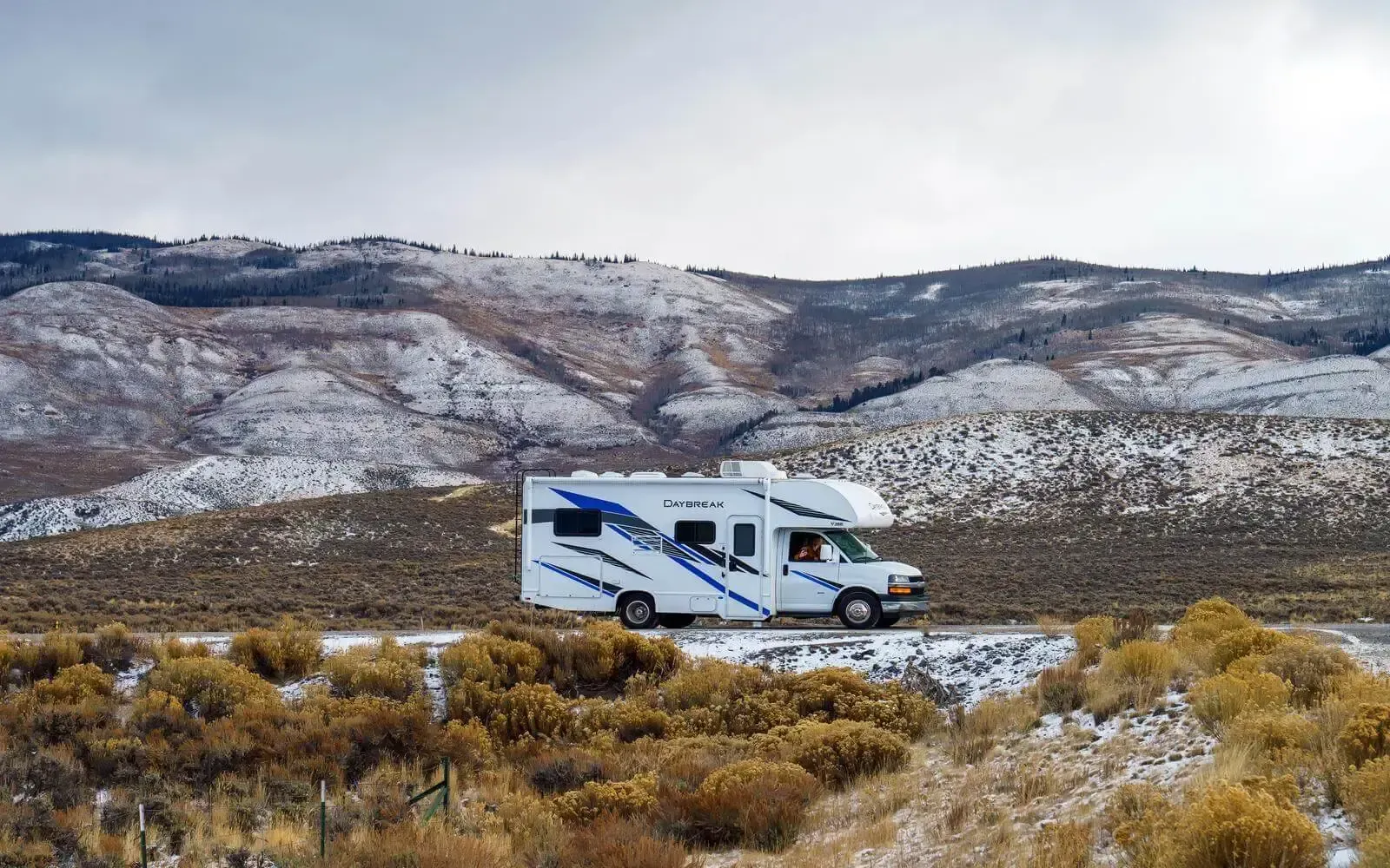 An RV driving on the road with snow on the ground and hills in the background.