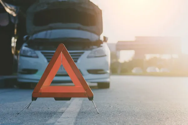 An orange emergency safety triangle on the side of the road with a car with its hood up in the background.