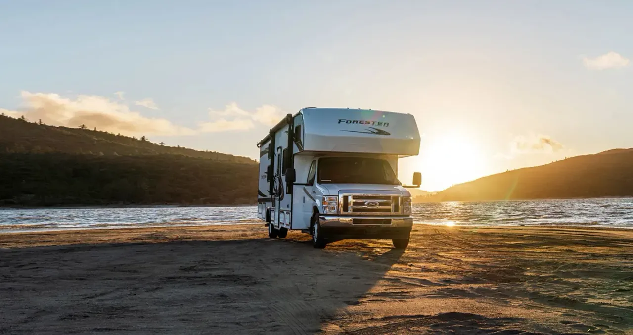 An rv parked in front of a body of water with the sun setting behind two mountains in the background.