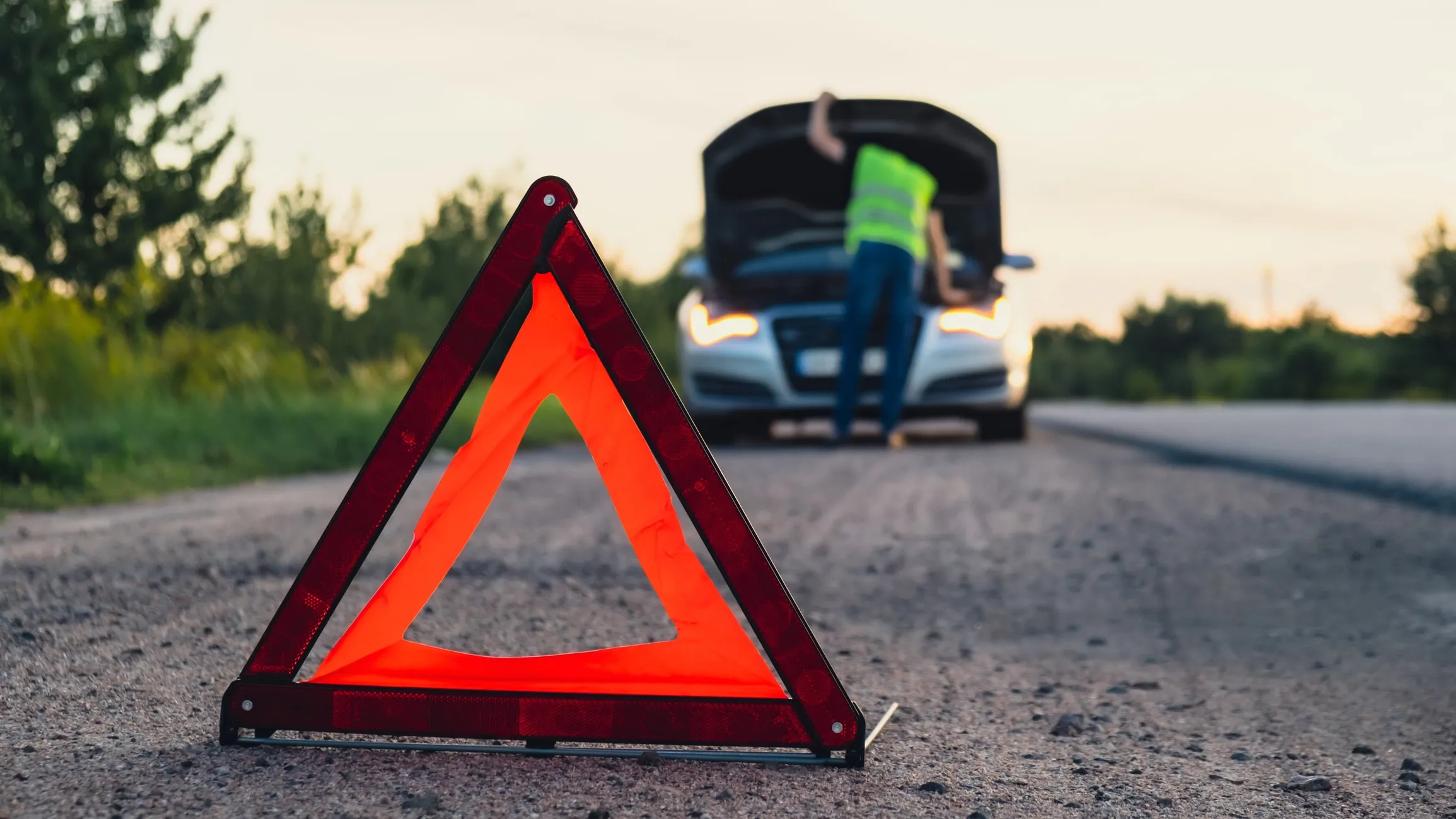 An orange emergency triangle on the road with a broken-down car and a person in a safety vest looking under the hood.