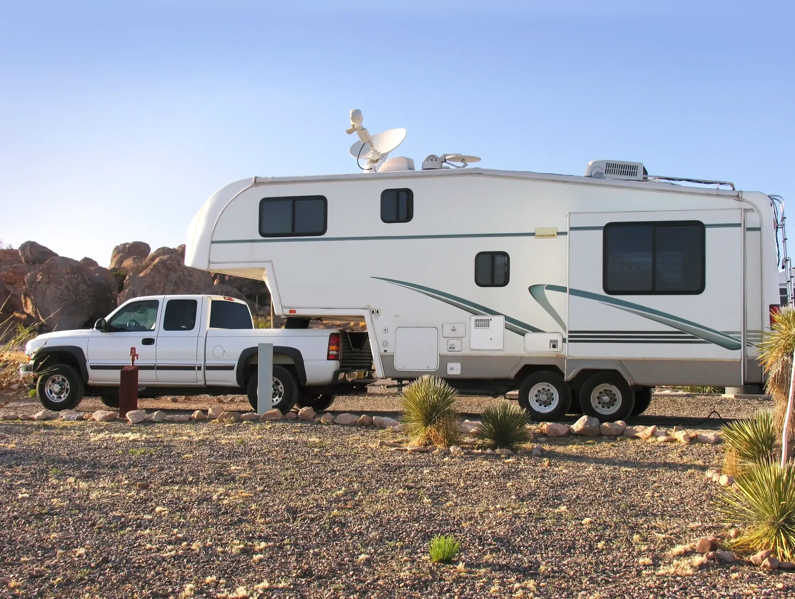 A white pick up truck pulling a fifth wheel camper on a gravel road with cactuses background. 