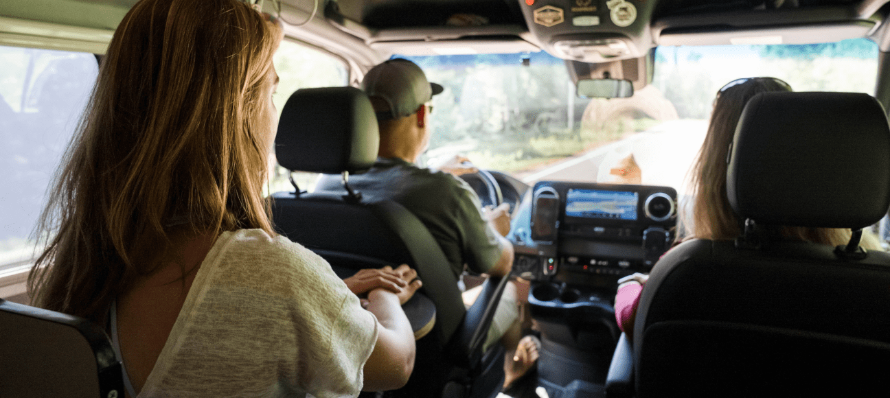 Three people ride in a van driving down a road on a sunny day.