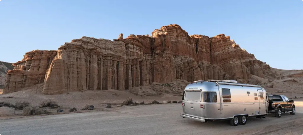 A truck tows a silver Airstream trailer in front of a towering red sandstone rock formation