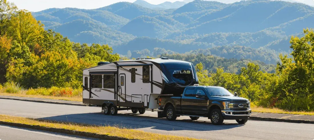 A truck tows an Atlas travel trailer with lush green rolling mountains in the background.