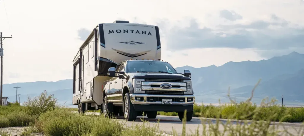 A truck tows a Montana fifth week along a road in with a backdrop of tall rocky mountains.