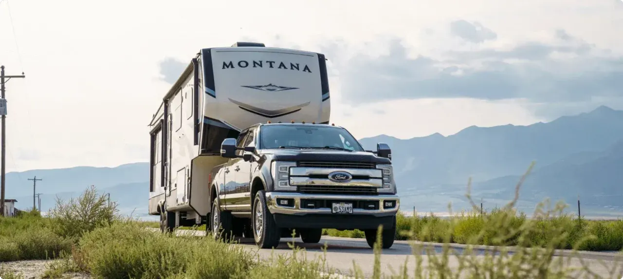 A truck tows a Montana fifth week along a road in with a backdrop of tall rocky mountains.