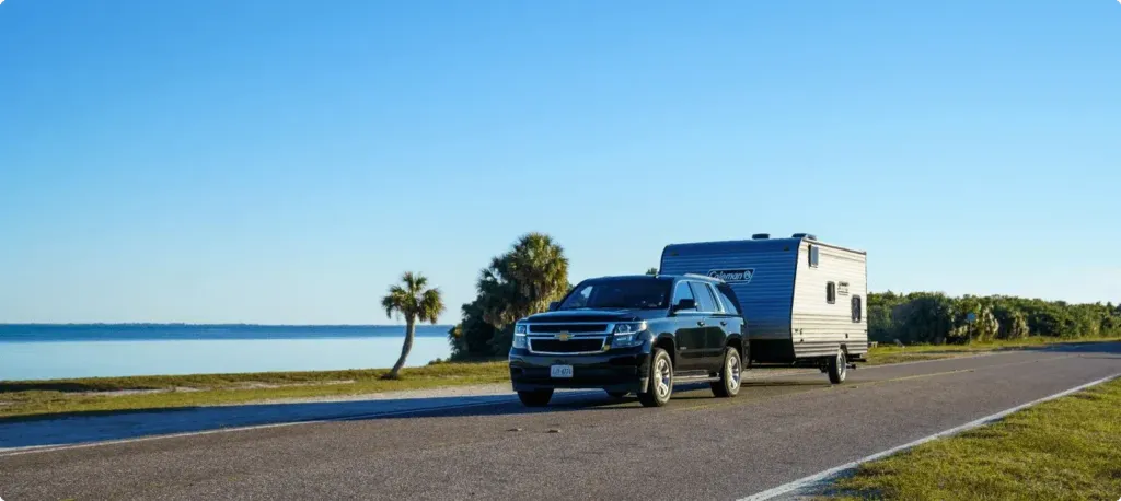 An SUV pulls a trailer alongside a coastal waterway with a palm tree in the background.