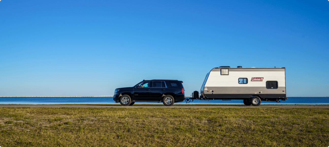 An SUV pulls a Coleman trailer along a road with a coastal waterway in the background.