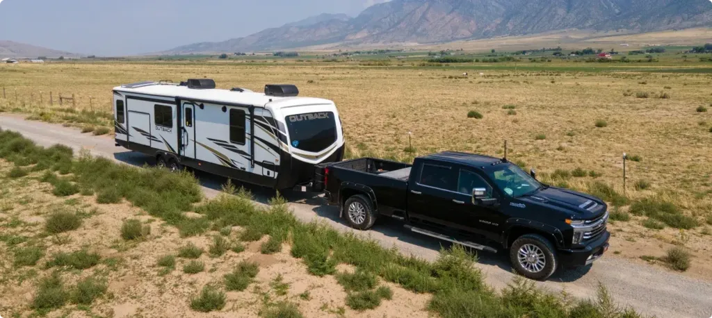 A truck tows an Outback fifth wheel down a gravel road surrounded by a dusty field with mountains in the background