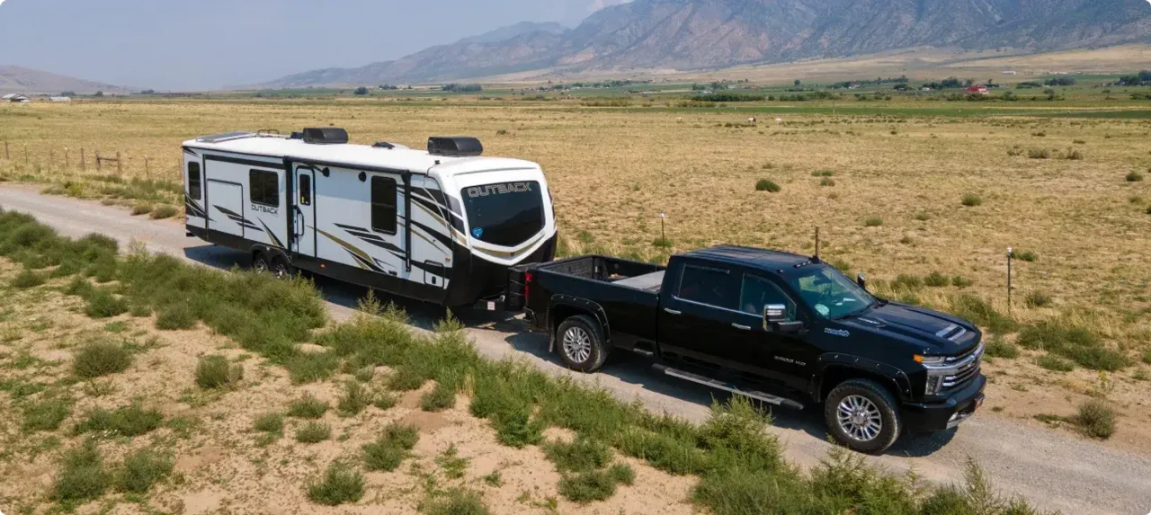 A truck tows an Outback fifth wheel down a gravel road surrounded by a dusty field with mountains in the background