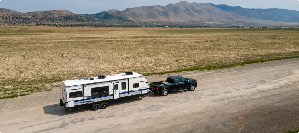 A truck tows a fifth wheel in front of a wide dusty field with tall rocky mountains in the background