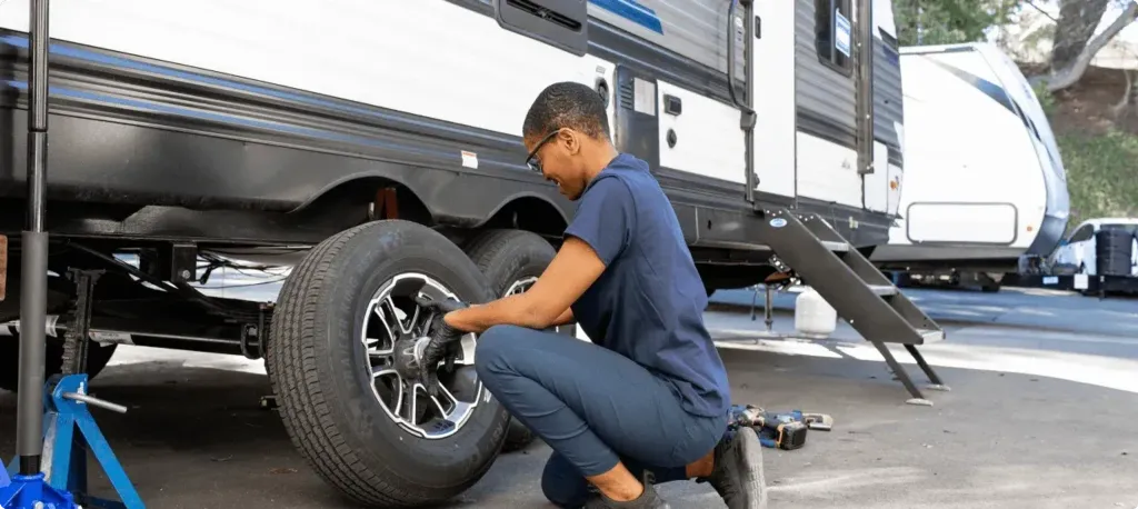 A woman kneels on the ground replacing a tire on her travel trailer. 