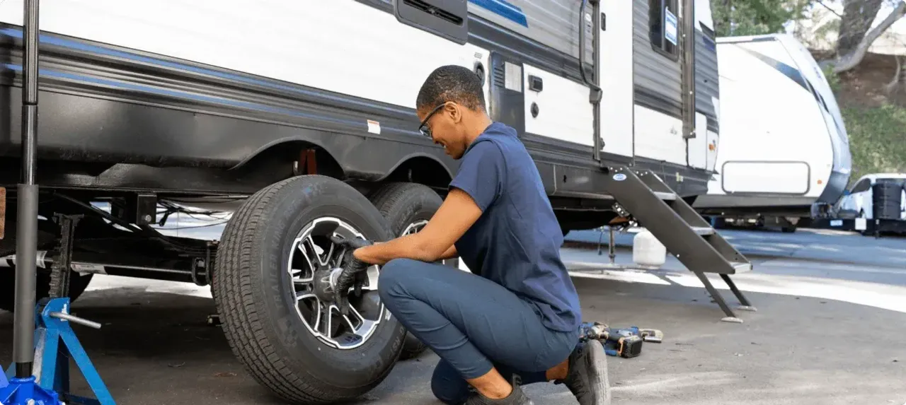 A woman kneels on the ground replacing a tire on her travel trailer. 