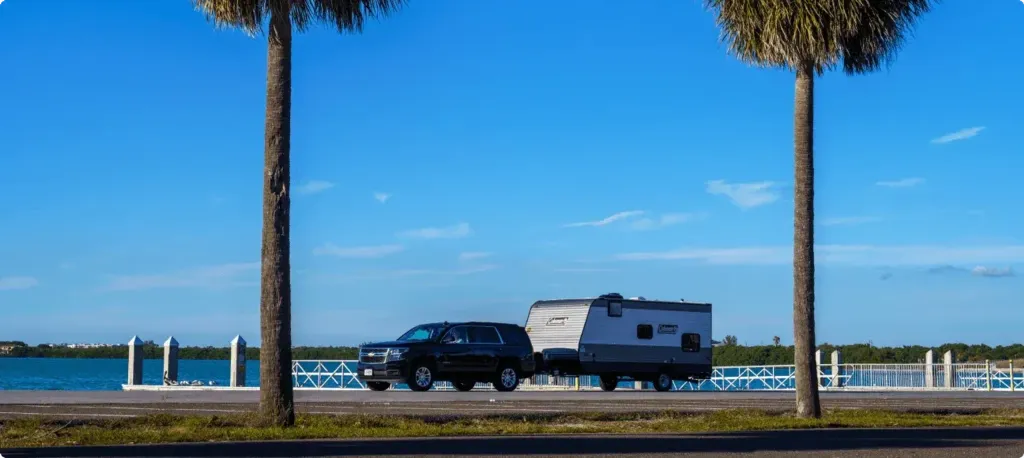 An SUV pulls a Coleman trailer along a road with a coastal waterway in the background.