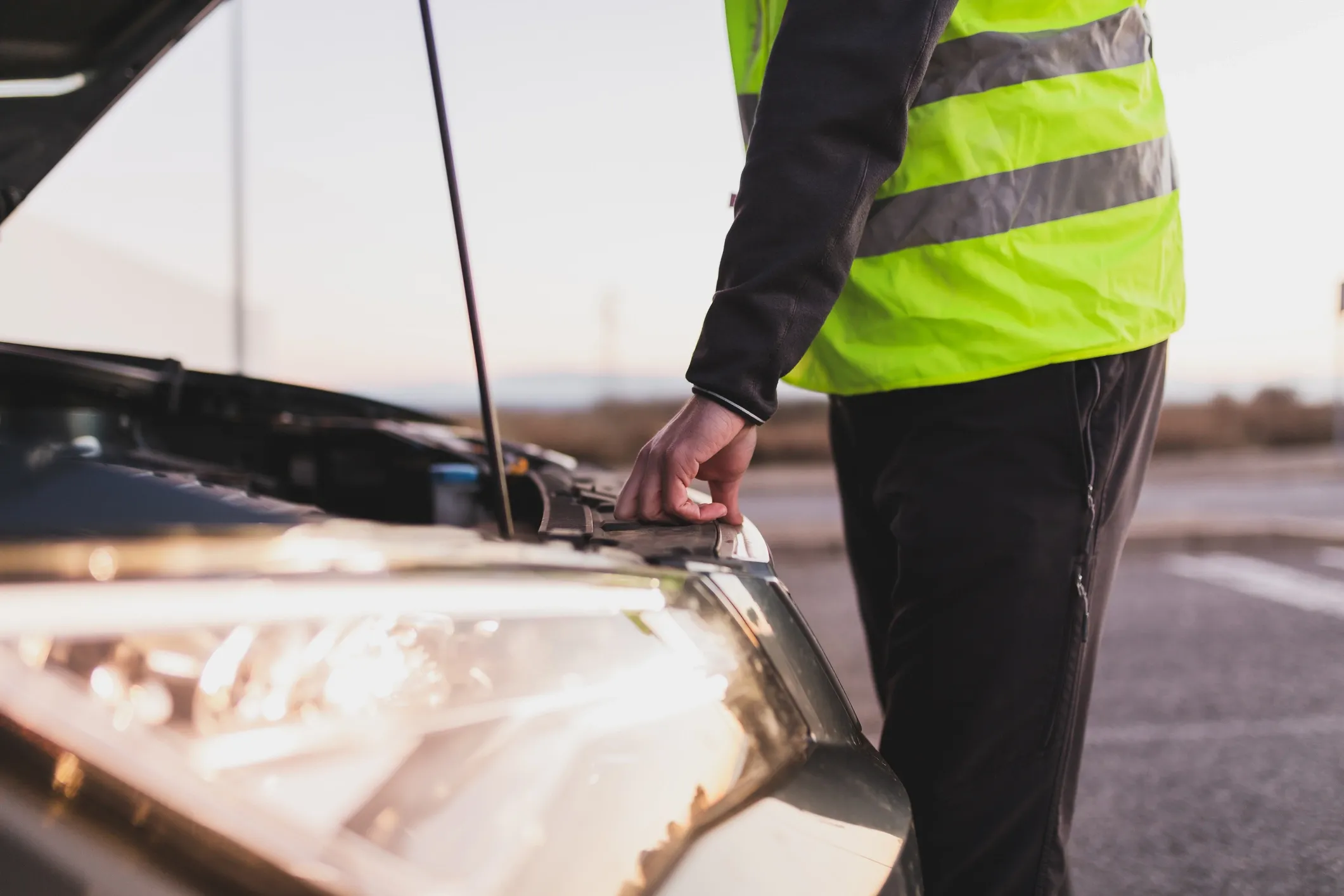 A roadside assistance technician in a high visibility safety vest looking underneath the hood of a car.