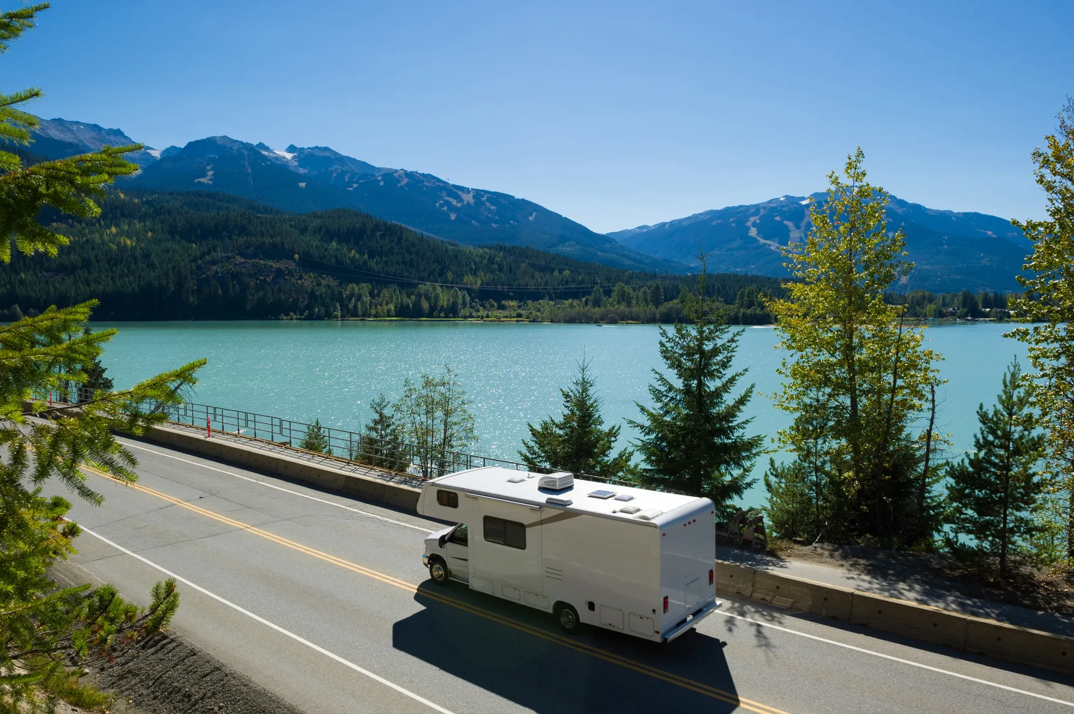 An RV driving on the road with a large lake next to the road and mountains in the distance. 