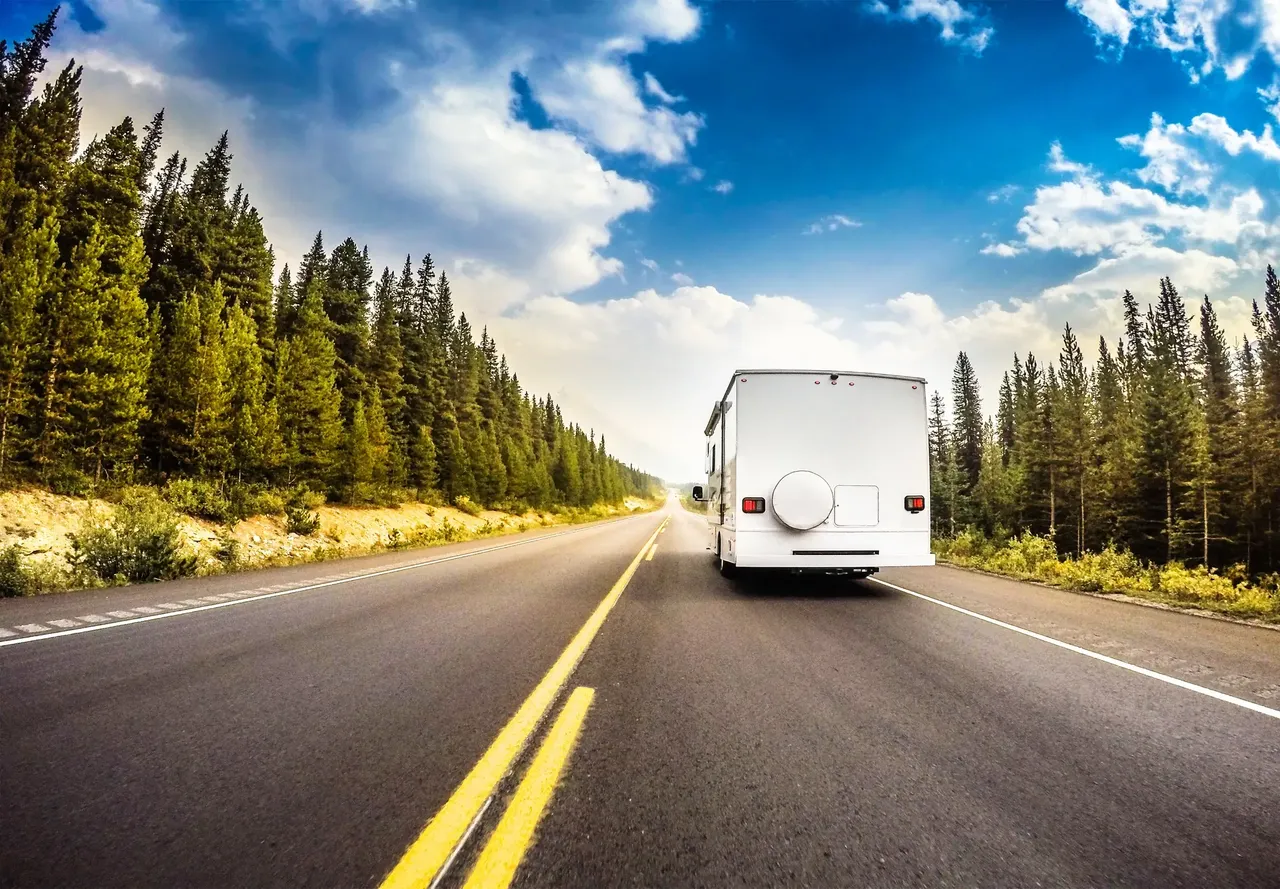 A white RV driving away on the right side of a road with a blue sky, white clouds, and evergreen trees on both sides of the road.