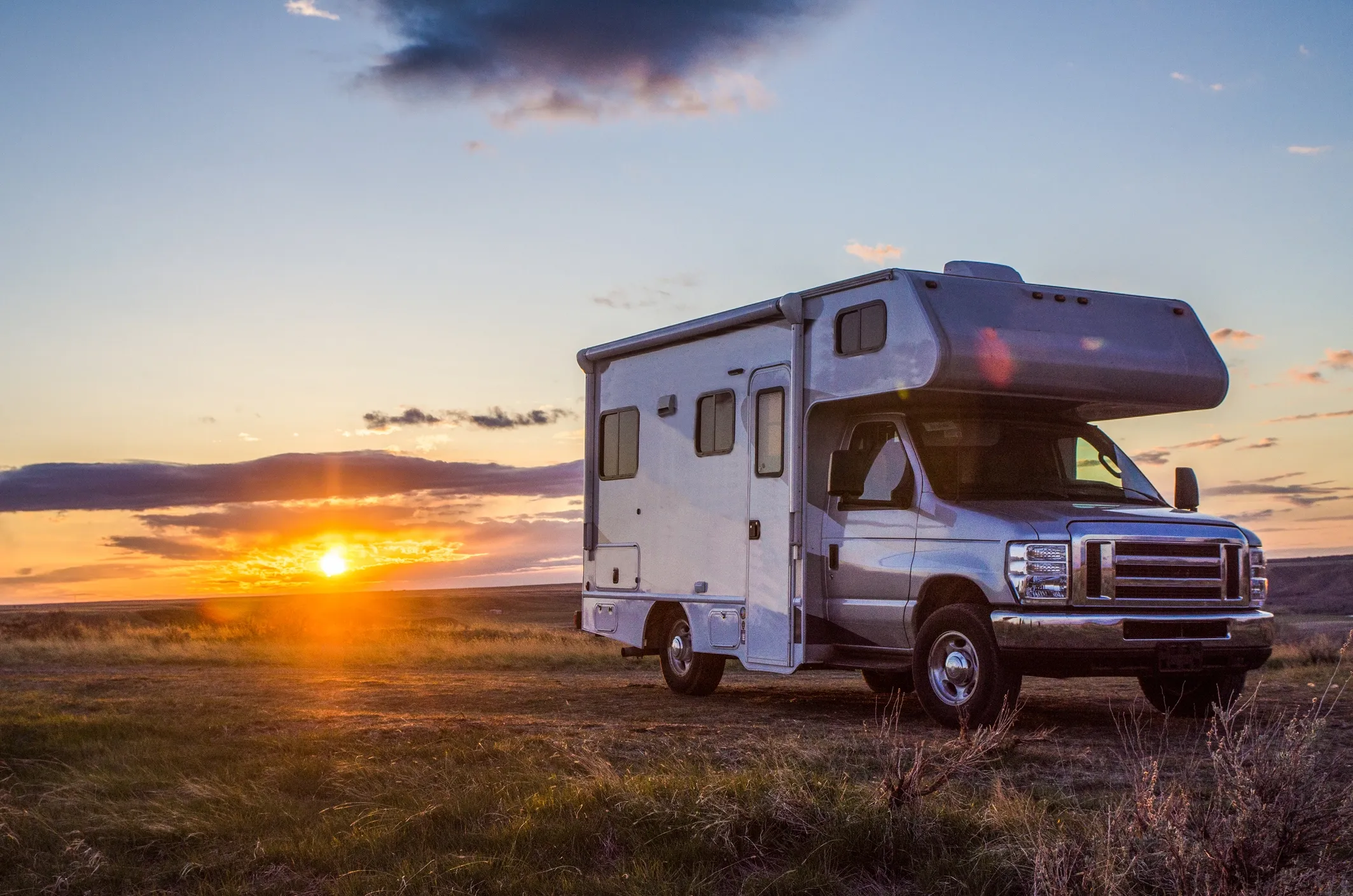 An RV parked in a grassy field with the sun rising out in the distance. 